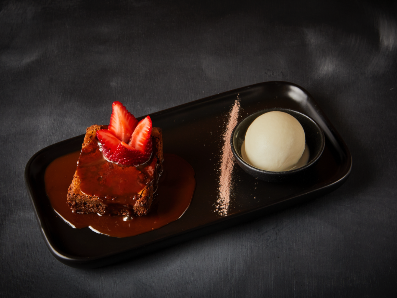 Brownie with strawberries, chocolate sauce, and ice cream on a black plate.