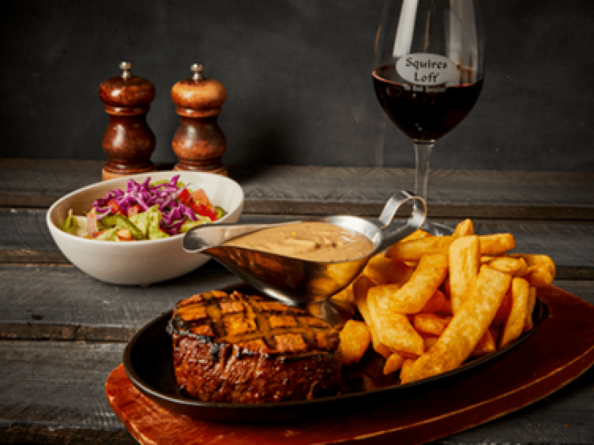 Steak, fries, salad, sauce, and red wine on wooden table with pepper mills.
