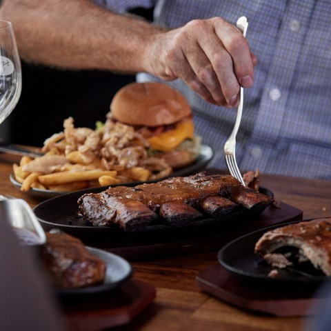 Person cutting ribs with fork at table, burger and fries in background.