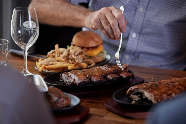 Person cutting ribs with fork at table, burger and fries in background.