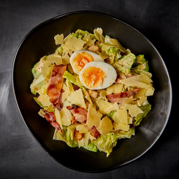 Plate of Caesar salad with bacon, boiled egg, and shaved Parmesan on a dark background.