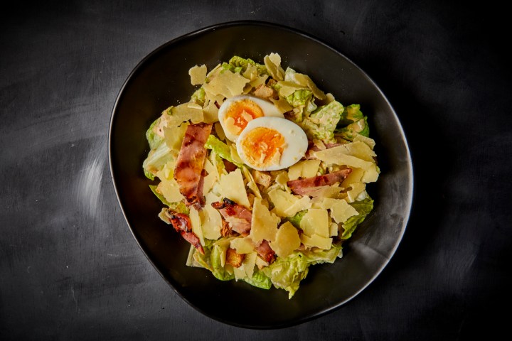 Plate of Caesar salad with bacon, boiled egg, and shaved Parmesan on a dark background.